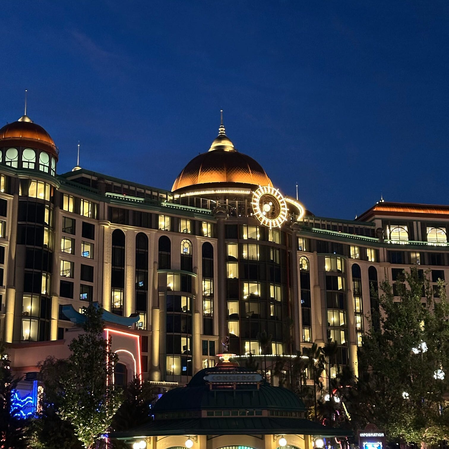 Night view of a beautifully lit building featuring a prominent dome and decorative clock, with surrounding trees and structures illuminated.
