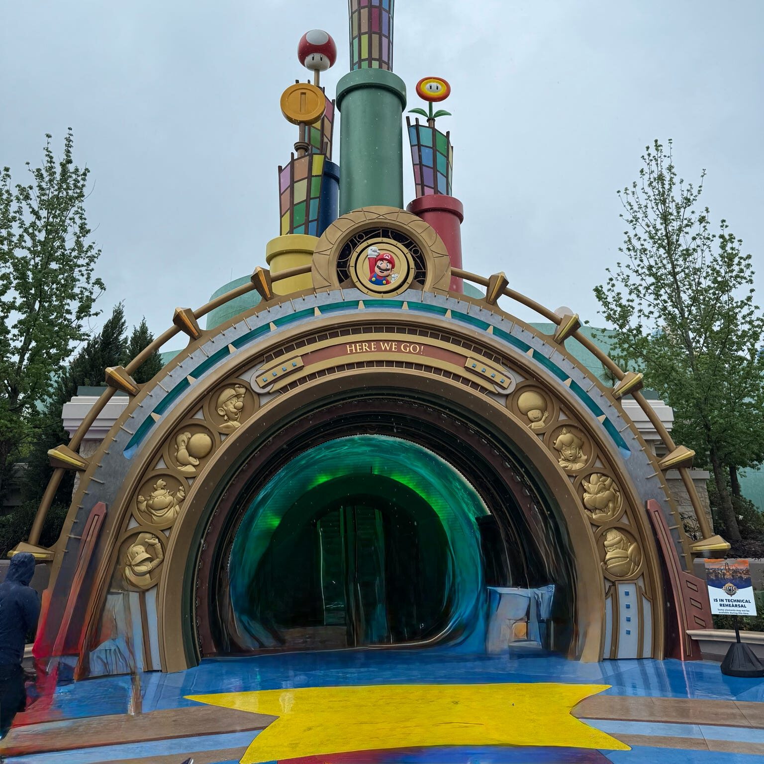 Entrance to Super Nintendo World featuring colorful structures and a large tunnel labeled 'HERE WE GO'.